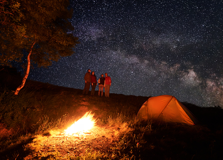Group of hikers enjoying the unusual sky strewn with bright stars during the night camping. In the foreground, a fire burns near the tent and the trees. Travel recreational outdoor activity concept.の写真素材
