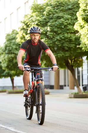 Handsome man bicyclist in cycling garment and protective helmet, looking at camera, riding bike down empty city center alley surrounded by green trees. Sportsman training outdoors improving skills.の写真素材