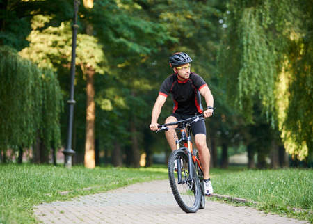 Young man cyclist in professional garment looking to side while riding bicycle down park alley on summer day. Sportsman training thinking about future win in contest. Exercising, reaching goalの写真素材