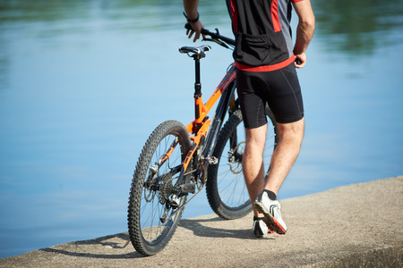 Back crop view of guy bicyclist wearing cycling clothes walking next to bicycle down pier to city lake. Sportsman resting and relaxing after training and exercising outdoors. Healthy lifestyle, natureの写真素材