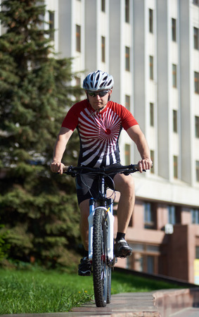 Young male bicyclist wearing professional cycling clothing and helmet riding bike, performing extreme tricks on street edging in front of blurred city building. Concept of healthy lifestyleの写真素材