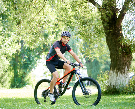 Sporty male biker in cycling sportswear and protective gear riding bike along green park alleys, smiling and looking at camera. Concept of healthy lifestyle, connecting with natureの写真素材