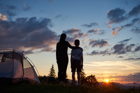 Rear view of the mother hugs her son near the tent on top of the mountain enjoying the view of the sunset, on the background - mountains and hills under incredibly beautiful evening sky with cloudsの写真素材