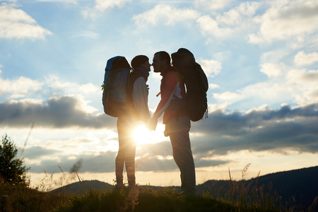 Pair of tourists in love with backpacks facing each other at sunset in the mountains with a landscape of mountains and cloudy skyの写真素材