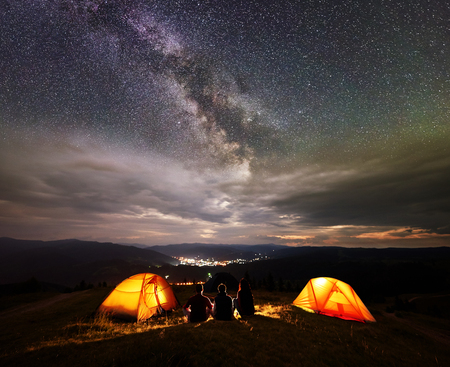 Rear view silhouette three tourists having a rest together beside camp, campfire and two orange tents at night on the top of mountain under wonderfull starry sky with many stars on backgroundの写真素材