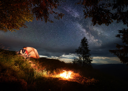 Couple backpackers in love sitting on a hill near the glowing orange tent, enjoying a burning fire under the night sky with bright stars and milky way. Powerful mountains and hills in the distanceの写真素材