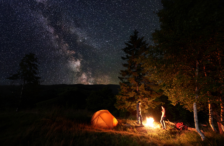 Guy and girl backpackers standing by the campfire near the woods and illuminated orange tent under the starry sky which is clearly visible milky way. Night camping near forest in the mountainsの写真素材
