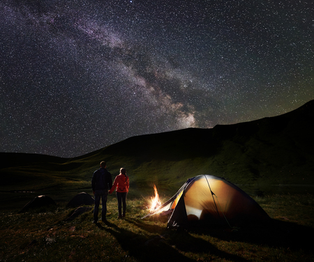 Back view of male and female backpackers holding hands, enjoying milky way on the starry sky at night camping. Beside illuminated tent and campfire is burning. Unusual landscape of mountains and lakeの写真素材