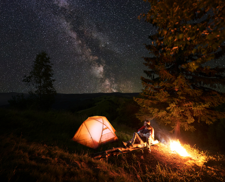Romantic couple hikers sitting on log beside illuminated orange tent, hugging and admire scenery of bright starry sky and Milky way. Pair having a rest by campfire at camping near forest in mountainsの写真素材