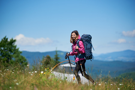 Smiling woman backpacker with backpack and trekking sticks near tent, wearing sports clothes, walking on the top of a hill against blue sky, enjoying sunny day in the mountains.の写真素材