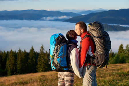 Close-up of a man kisses a woman in the sun at sunset against the background of the forest, mountains and thick haze above them. The concept of outdoor activities in the mountainsの写真素材