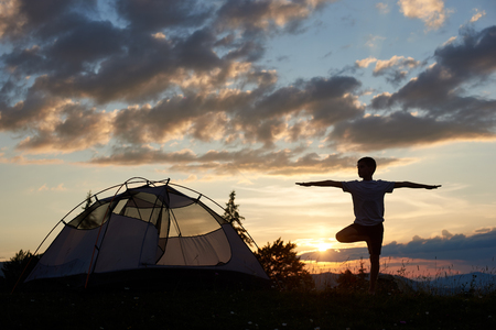 Silhouette of boy at practice yoga on the top of mountain at the dawn near campsite under sky with clouds and bright morning sun. Breathtaking landscape of mountains and hills above which sun risesの写真素材