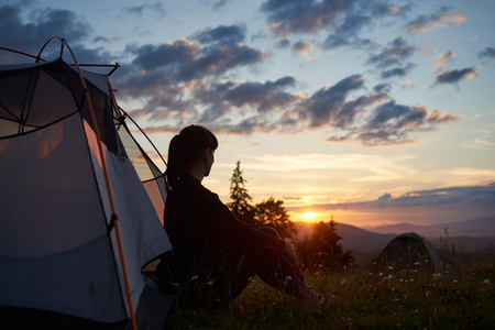 Woman tourist sitting in tent at the top of mountain enjoying the sunset on blurred background of mountains and hills under incredible beautiful evening sky with clouds. Concept of healthy lifestyle.の写真素材