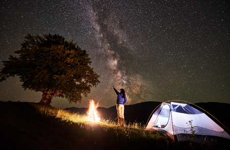 Back view woman tourist having a rest at night camping in mountains. Female hiker standing beside campfire and illuminated tent near big tree, pointing at incredible beautiful starry sky and Milky wayの写真素材