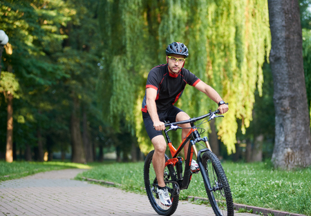 Sporty guy bicyclist in cycling clothes and protective helmet looking to camera while riding bike down park alley on summer day. Exercising, reaching goalの写真素材