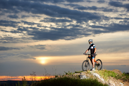 Cyclist with a bicycle rests on a cliff top in the mountains enjoying the sun at sunset under an unusual sky with beautiful clouds. Man in sports clothes and helmetの写真素材