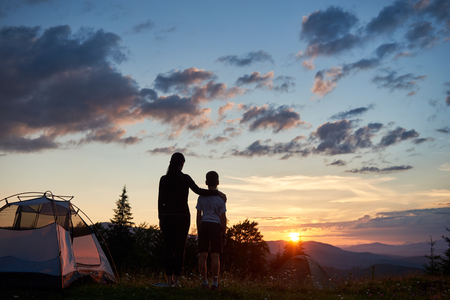 Back view silhouettes of woman hugging child near a camping in the mountains at dawn standing on the grass with wildflowers. The family enjoys the rising sun above the mountains and hillsの写真素材