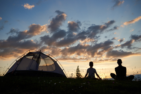 Rear view of a silhouette of woman and boy sitting on the grass with wild flowers relaxing near campsite at sunset. The family enjoys the scenery of the mountains and hills in evening lightの写真素材