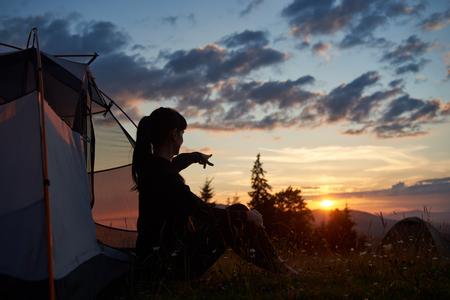 Attractive woman sits in a profile near a tent on the grass with wildflowers and shows her hand in the distance to the rising sun over the mountains. A fascinating landscape of the dawn in mountainsの写真素材