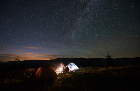Back view of mother and two children tourists having a rest at night camping in mountains, sitting on log beside campfire and glowing tent, looking at beautiful starry sky and Milky way in eveningの写真素材