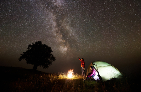 Night camping near silhouette of big tree on grassy valley. Tourist man standing near burning campfire, pointing at beautiful starry sky and Milky way to young woman sitting in glowing tent entranceの写真素材