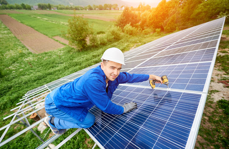Construction worker connects photo voltaic panel to solar system using screwdriver. Professional installing and construction of solar system, smiling to the camera. Alternative energy conceptの写真素材