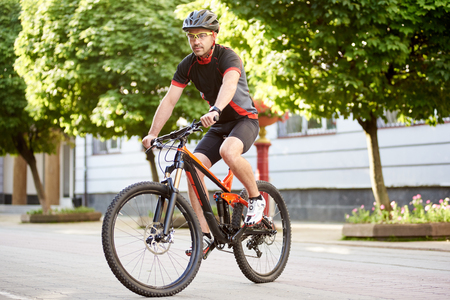 Athletic guy cyclist in cycling clothes and protective helmet, riding bike down empty city center alley surrounded by green trees. Sportsman training outdoors improving skills.の写真素材