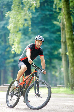 Athletic man bicyclist in professional garment and helmet riding bicycle park alley, looking at camera, smiling. Happy sportsman training outdoors connecting with nature. Concept of healthy lifestyleの写真素材