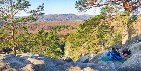 Beautiful sunny morning in mountains. Young tourist couple, bearded man and long-haired girl sitting in sleeping bags on big rocks enjoying peace and quietness. Tourism and traveling concept.の写真素材