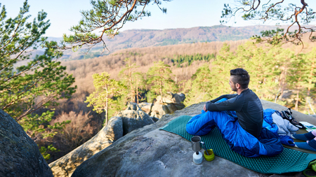 Young tourist, bearded handsome man sitting alone in sleeping bag on big rock having coffee and enjoying beautiful mountain view on clear sunny summer morning. Tourism and active lifestyle concept.の写真素材
