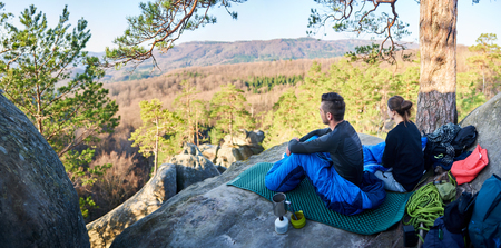 Young tourist travelers, handsome man and pretty girl camping on big rocks enjoying magnificent peaceful view of mountains covered with forest on early morning. Tourism and active lifestyle concept.の写真素材