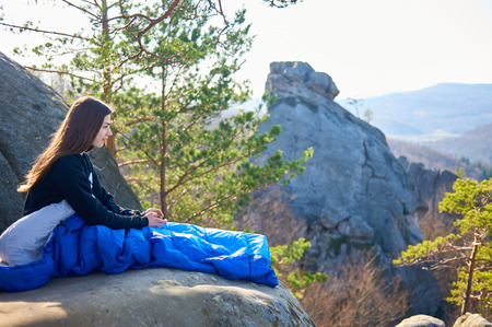 Beautiful young long-haired woman sitting alone in sleeping bag on big mountain rock and smiling dreamily enjoying fantastic view on clear sunny summer morning. Tourism and active lifestyle concept.の写真素材