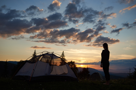 Rear view silhouette of young lady standing on top of a mountain near a camping at dawn enjoying the view of the mountains and hills and the rising sun above them with a beautiful sky in the clouds.の写真素材