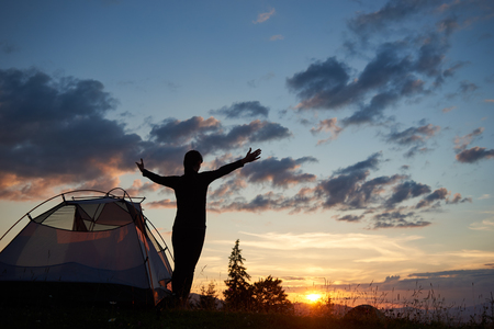 Rear view silhouette of female backpacker standing with open arms near a camping in the mountains at sunset under the evening yellow skyの写真素材