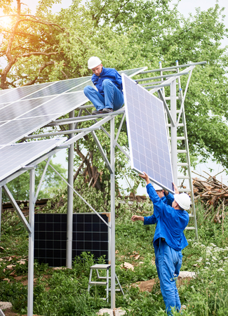 Installing of stand-alone solar photo voltaic panel system. Workers in hard-hats and blue overall lifting the solar module on metal platform. Alternative energy and professional construction concept.の写真素材