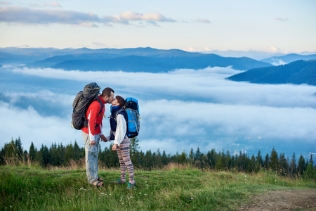 Romantic kiss on the background of mighty mountains in the fog under the sky with clouds. Male and female with backpacks holding hands on a green hilltopの写真素材