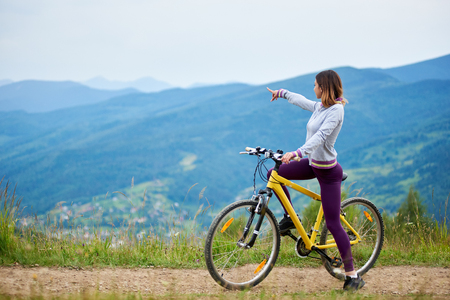 Sporty female cyclist riding on yellow bicycle on a rural trail, pointing at something in the distance. Mountains on the blurred background. Outdoor sport activity. Copy spaceの写真素材