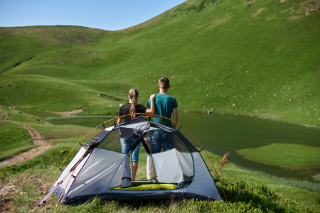 Rear view two young tourists enjoying lake near camping in the mountains covered greenery. Woman put her hand on the shoulder of the man. The concept of active outdoor resting at campingの写真素材