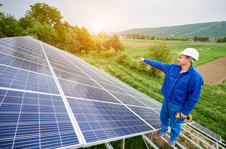 Construction worker with screwdriver standing on metal frame of photo voltaic solar system pointing on shiny surface. Alternative energy, ecology protection and cheap electricity production concept.の写真素材
