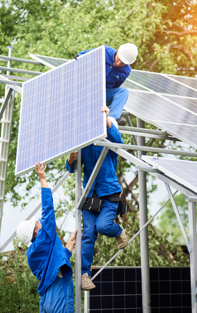 Installing of stand-alone solar photo voltaic panel system. Workers in hard-hats and blue overall lifting the solar module on metal platform. Alternative energy and professional construction concept.の写真素材