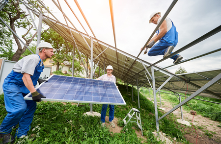 Installing of stand-alone solar photo voltaic panel system. Three technicians lifting big solar module on high metal platform. Investment in alternative energy and professional construction concept.の写真素材