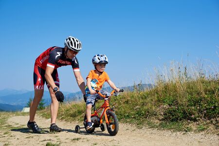 Verkhovyna, Ukraine - August 19, 2017: Young athletic father in professional biker outfit teaches small preschool boy to ride a bike on dusty empty mountain road. Travel and family recreation concept.のeditorial素材