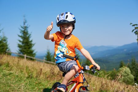 Verkhovyna, Ukraine - August 19, 2017: Small preschool boy in helmet cycling three-wheeled child bike down steep mountain hill shows thumb-up sign smiling in camera. Success and confidence concept.のeditorial素材