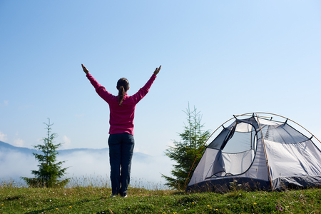 Back view of happy backpacker girl standing with raised arms on blooming hill at tourist tent under beautiful clear blue sky on bright summer morning. Tourism and traveling concept.の写真素材