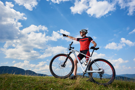 Professional male cyclist in sportswear and helmet standing with cross country bike on top of hill, pointing to the blue sky with clouds on summer sunny day. Active lifestyle and outdoor sport conceptの写真素材