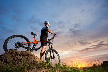 Cyclist standing alone with orange bicycle and holding hands on handle bar. Athletic man wearing black sportswear and helmet and posing on hill. Concept of healthy lifestyle and sport activities.の写真素材