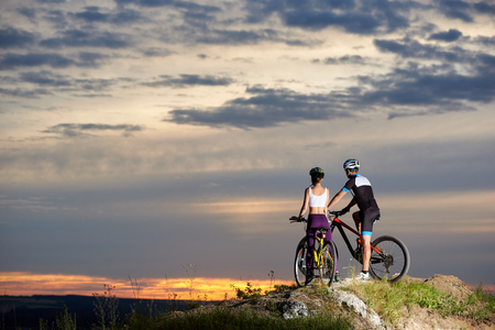 Back view of a couple riding a mountain bike on top enjoying the sunset and a beautiful evening in the mountains. Young people dressed in sporting clothes and helmetsの写真素材