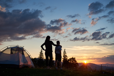 Rear view of silhouette mother hiker hugging her son in the camping on top of the mountain. Tourists enjoying view of sunset. Mountains and hills under amazing evening sky with clouds on backgroundの写真素材