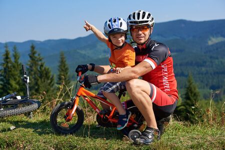 Verkhovyna, Ukraine - August 19, 2017: Happy tourist family: athletic biker father lovingly holds his preschool son sitting on child bicycle with thumb-up gesture on blue foggy mountains background.のeditorial素材