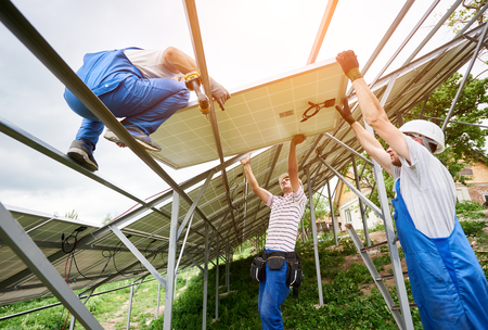 Installing of solar photo voltaic panel system. Three technicians lifting heavy solar module on high platform. Investment in alternative energy, money saving and professional construction concept.の写真素材